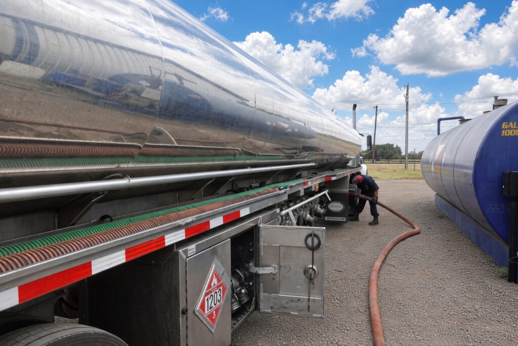 A worker connects a long hose to a shiny tanker for fuel transfer, with a blue storage tank visible nearby under a cloudy sky.