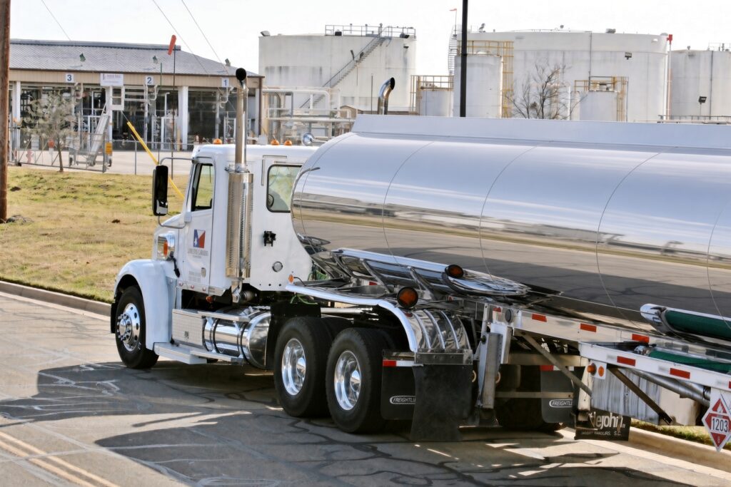 A shiny, silver tanker truck parked near industrial facilities, showcasing its chrome detailing and warning symbols. It represents logistics in fuel transport.