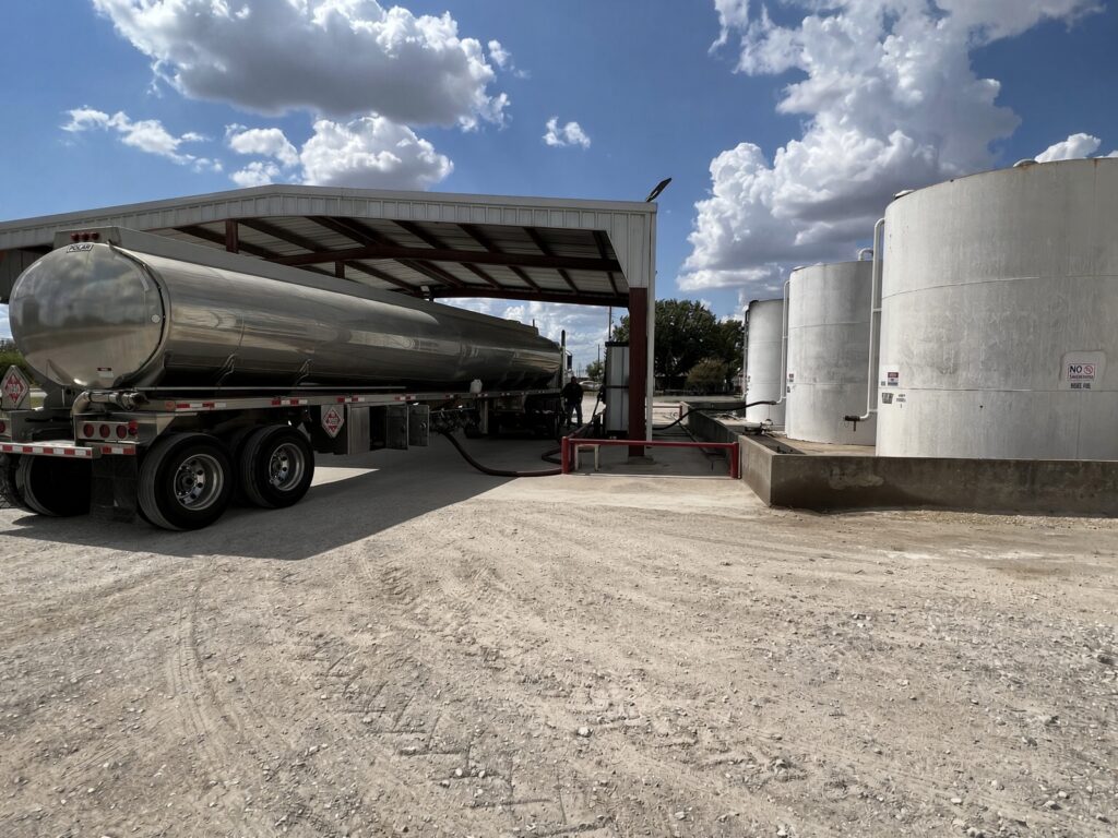 A stainless steel tanker is parked at a facility, unloading liquid into large storage tanks under a covered structure with blue skies above.